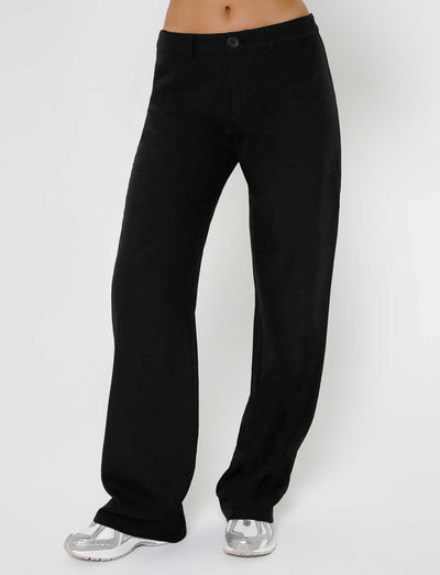 A person standing in front of a plain white background wears the MARJORIE PANT - BLACK from TIGERMIST, featuring black, wide-legged, high-waisted pants with a single button at the waistband, paired with white sneakers.