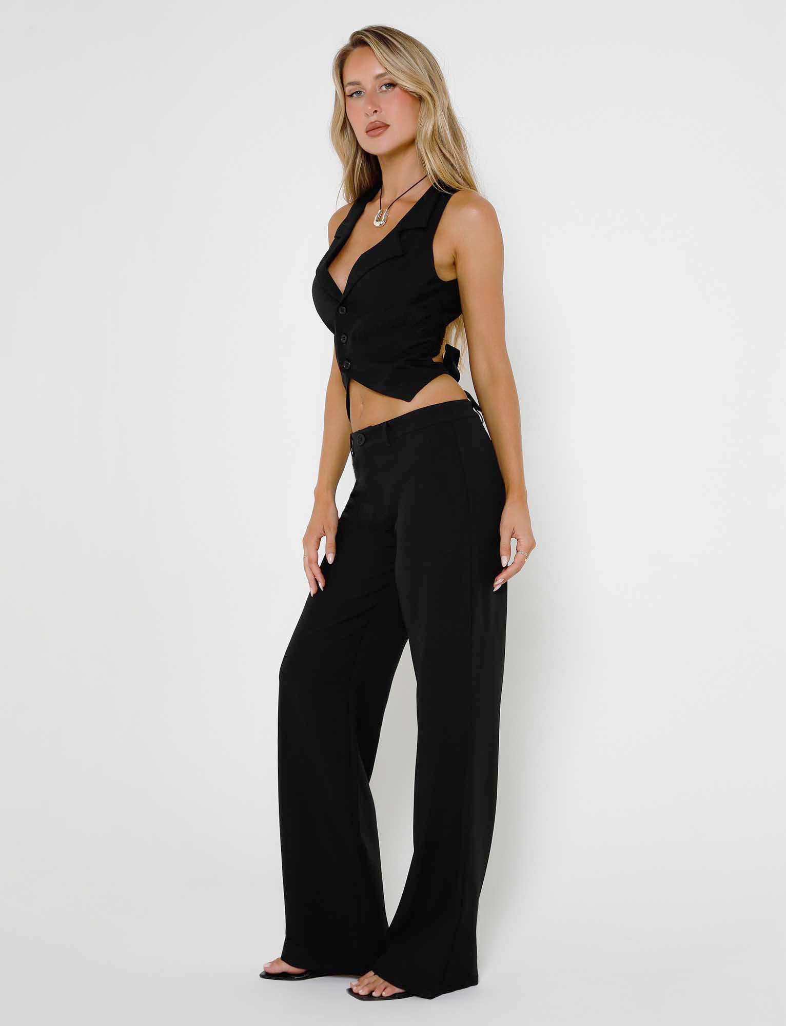 A woman stands confidently against a plain white background, dressed in a chic, black sleeveless vest and matching high-waisted MARJORIE PANTS from TIGERMIST. Her blonde hair is styled in loose waves, and she accessorizes with a necklace and open-toe sandals.