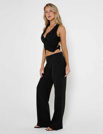 A woman stands confidently against a plain white background, dressed in a chic, black sleeveless vest and matching high-waisted MARJORIE PANTS from TIGERMIST. Her blonde hair is styled in loose waves, and she accessorizes with a necklace and open-toe sandals.