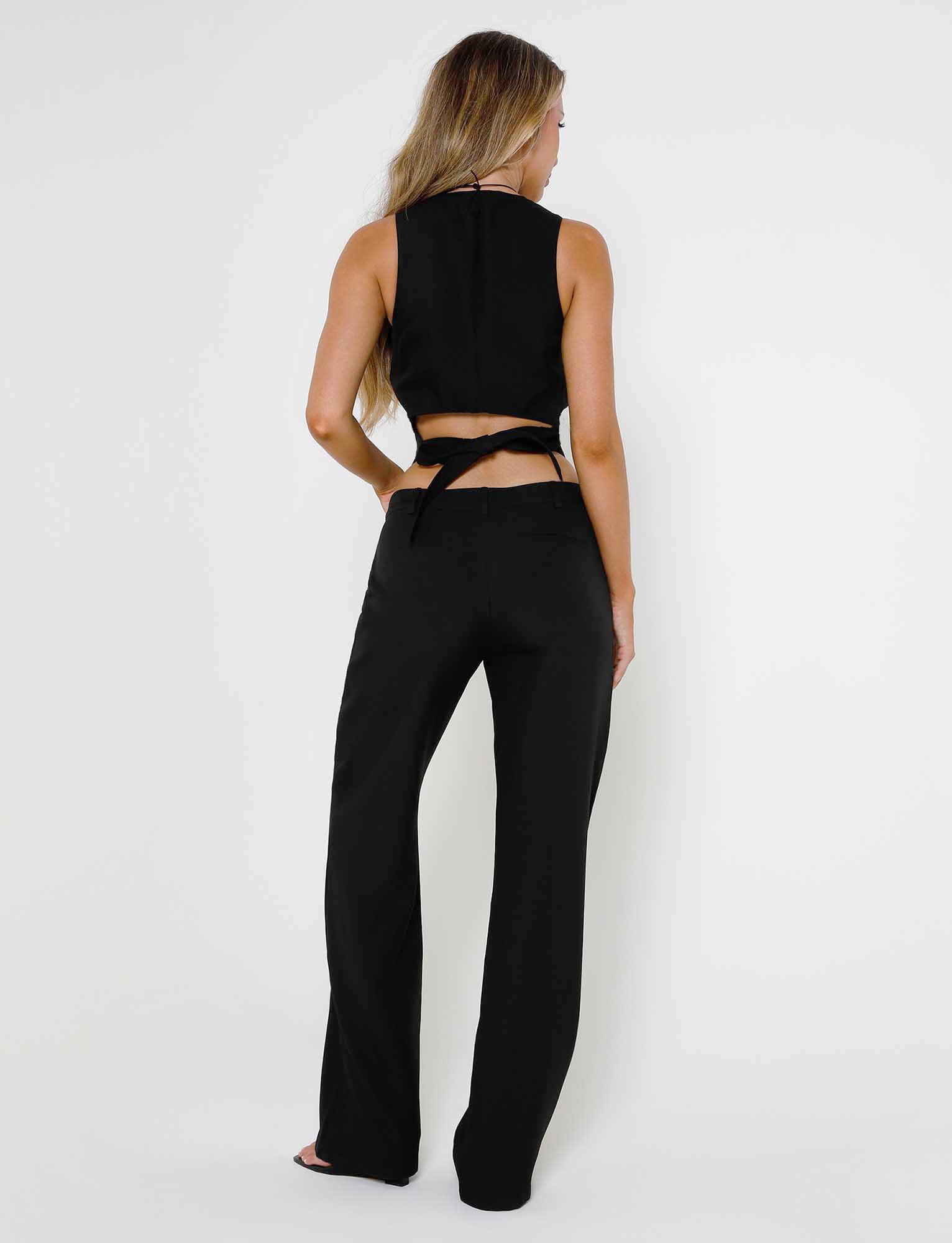 A woman with long, wavy hair stands facing away from the camera. She is wearing a sleeveless black crop top with a crisscross tie at the back and matching MARJORIE PANT - BLACK from TIGERMIST. The background is plain white.