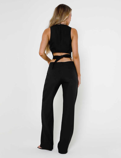 A woman with long, wavy hair stands facing away from the camera. She is wearing a sleeveless black crop top with a crisscross tie at the back and matching MARJORIE PANT - BLACK from TIGERMIST. The background is plain white.
