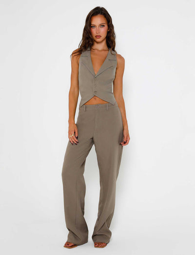 A woman with long brown hair stands against a plain white background, embodying the sleek elegance of TIGERMIST. She is wearing a sleeveless, button-up vest and the MARJORIE PANT in moss green khaki. Her left hand rests on her thigh while her right hand hangs by her side. She is expressionless, directly facing the camera.