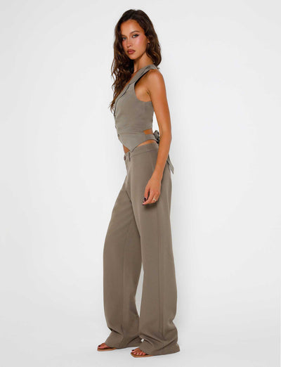 A woman stands against a plain white background wearing the MARJORIE PANT in moss green from the TIGERMIST collection. She has long, wavy brown hair and is looking directly at the camera with a neutral expression. She is barefoot and pairing her trousers with a gray sleeveless top featuring side cutouts.