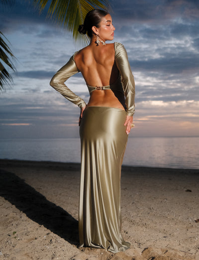 A woman poses on a sandy beach at sunset, facing away from the camera. She wears the Thessaly Skirt in Matcha Green by Tiger Mist, which drapes elegantly to the ground. Her hair is styled in an elegant updo, and she stands under a palm tree with the calm ocean and cloudy sky in the background.