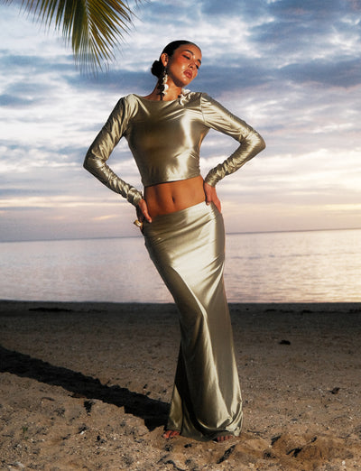 A woman stands on a sandy beach at sunset, wearing a shiny, long-sleeved silver crop top paired with the MATCHA GREEN THESSALY SKIRT from TIGERMIST. She poses with one hand on her hip and the other hanging by her side. The ocean and a partially cloudy sky appear in the background.