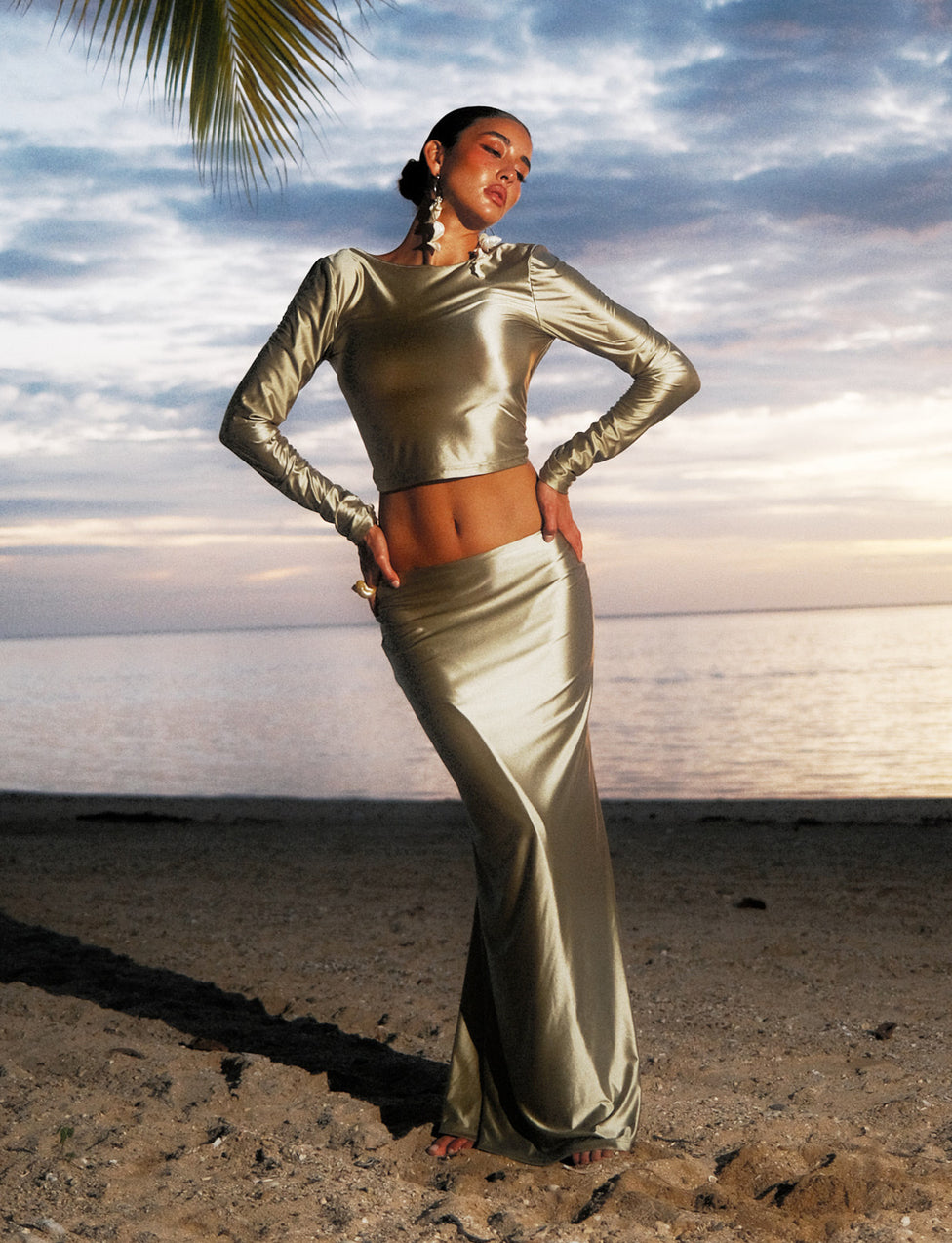 A person poses on a sandy beach at sunset, wearing a shiny TigerMist Thessaly Long-Sleeved Crop Top in matcha green and matching long skirt. The ocean and cloudy sky create a serene backdrop, with a palm tree branch visible in the upper left corner.