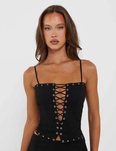 A woman with long brown hair is wearing the KERI TOP - BLACK from TIGERMIST, a sleek black sleeveless top featuring a lace-up front with metal eyelets. The top has thin shoulder straps and a fitted silhouette. She is standing against a plain white background.