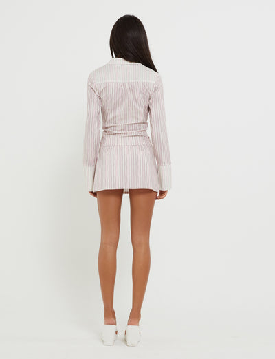 A person with long dark hair stands facing away from the camera. They are wearing the LILIOSA SKORT - PINK in pink pinstripe from TIGERMIST, paired with white heels against a plain white background.