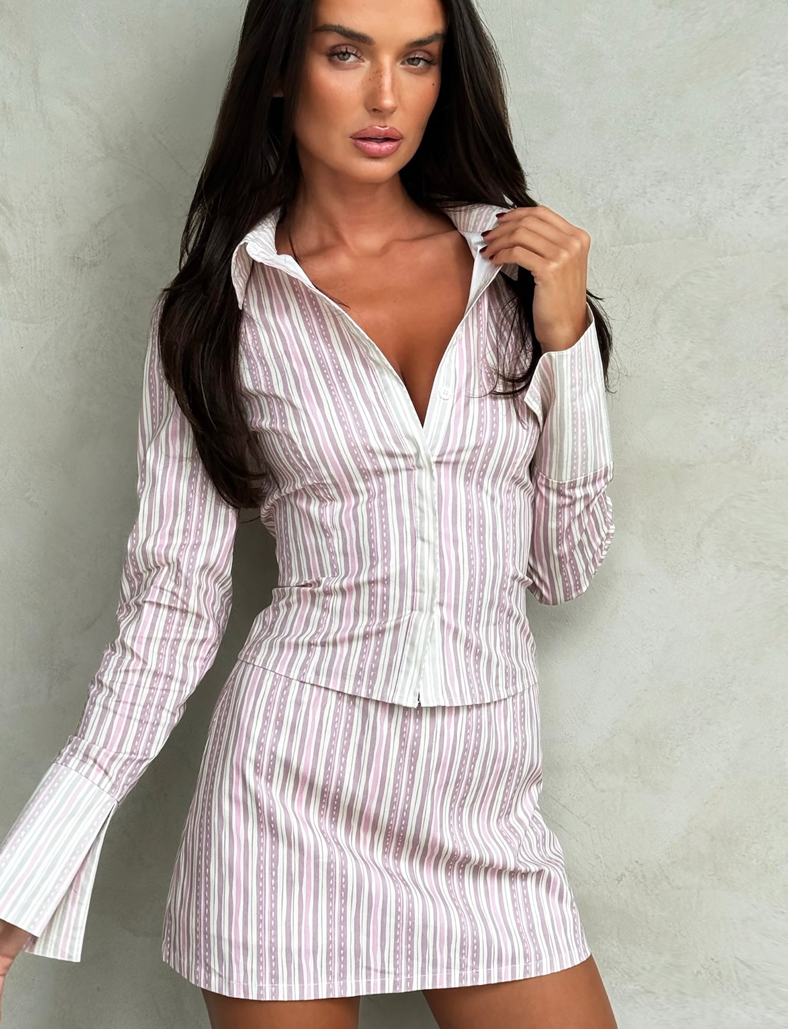 A woman with long dark hair stands against a textured light gray wall. She is wearing TIGERMIST’s LILIOSA SKORT in Pink Pinstripe, featuring a fitted blouse partially unbuttoned and a matching short skirt. She poses with one hand touching her hair and the other resting on her skirt.