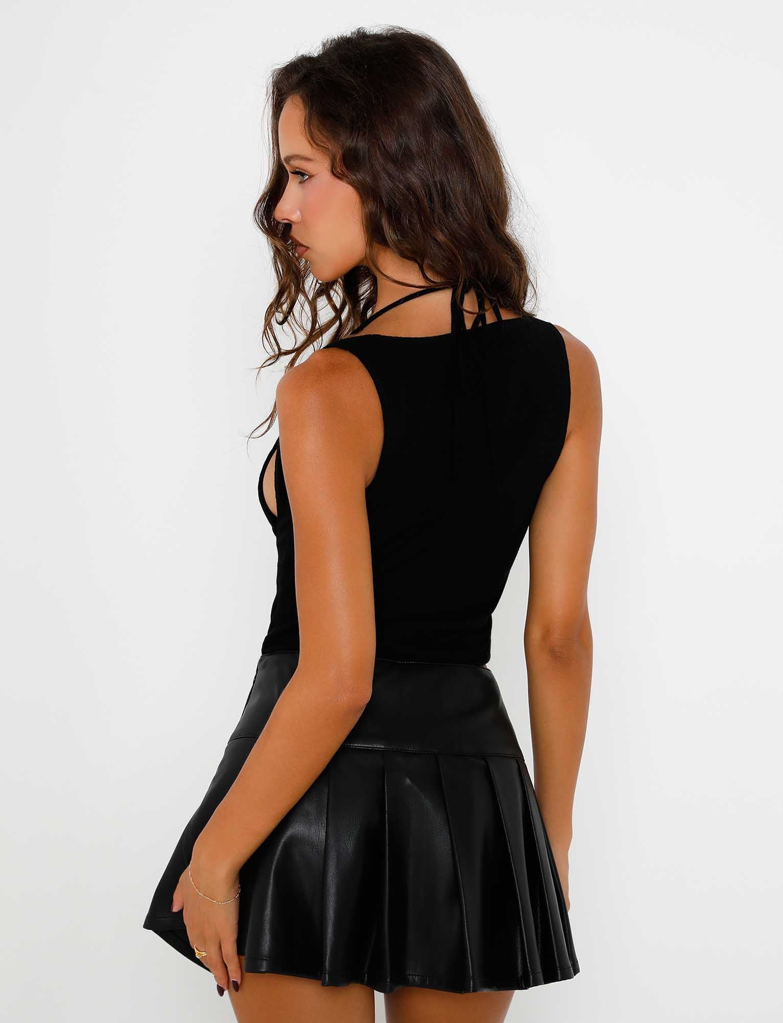 A woman with long wavy hair is shown from the back, wearing a black sleeveless halter top and the MERRI SKIRT - BLACK : BLACK PU by TIGERMIST. She has her left hand slightly raised, with her head turned to the left, exposing her profile. The background is plain white.