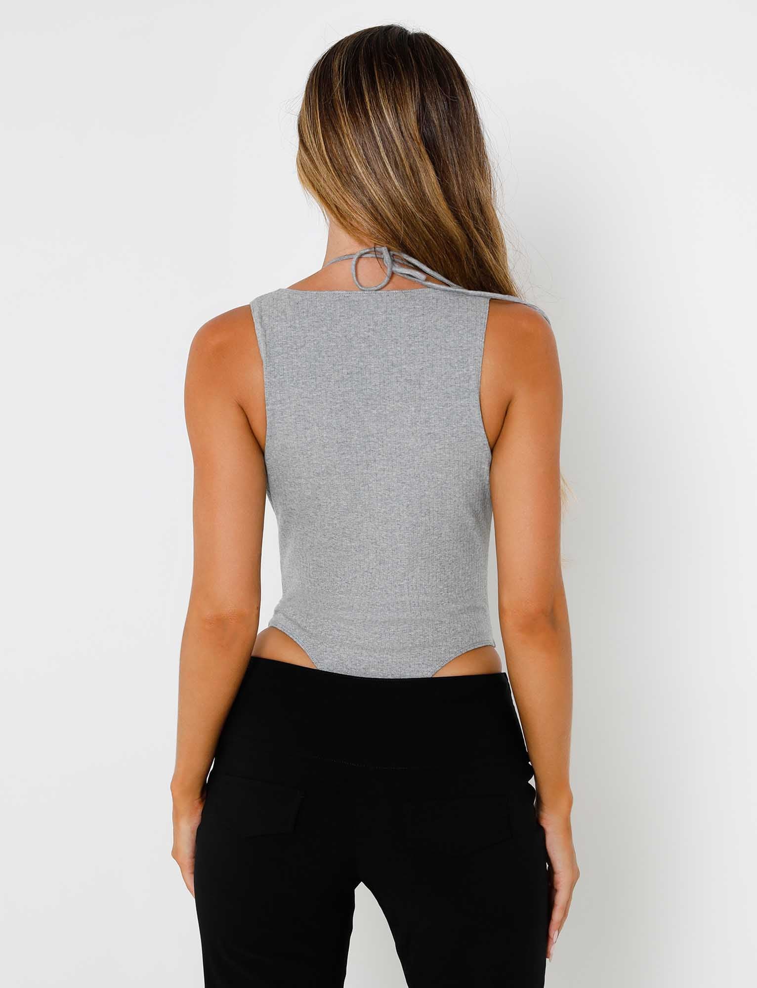 A woman with long, light brown hair is seen from the back, wearing the TARYN BODYSUIT - GREY and black pants from TIGERMIST. The halter top ties behind her neck, and her hair flows freely down her back. The background is plain white.