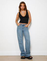 A woman stands against a white wall wearing the SACHA JEAN - MID BLUE by TIGERMIST, featuring a V-neck black top and black sandals. She has long hair, a necklace, and her hands rest in her pockets with the relaxed straight leg jeans giving an effortlessly stylish look.