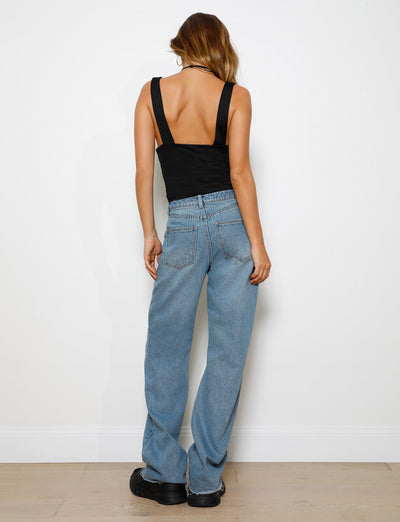 A person with long hair stands facing a white wall, wearing a sleeveless black top with an open back and SACHA JEAN - MID BLUE high-waisted jeans by TIGERMIST, featuring a raw hemline. They complete the look with relaxed straight leg jeans and black platform shoes.
