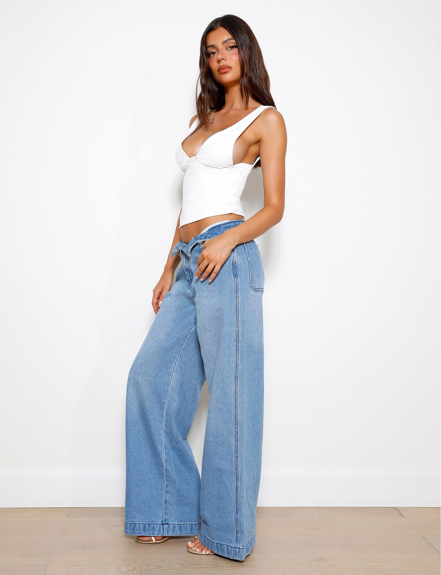 A woman poses against a white wall, wearing a white asymmetrical top and the TIGERMIST COHEN JEAN - DENIM, featuring a wide leg silhouette. She stands with one hand on her hip and her long hair down.