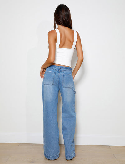 A person with long hair is facing a wall, dressed in a white sleeveless top and the TIGERMIST COHEN JEAN - DENIM featuring a wide leg silhouette. The room showcases a light-colored wooden floor and has a plain white wall in the background.