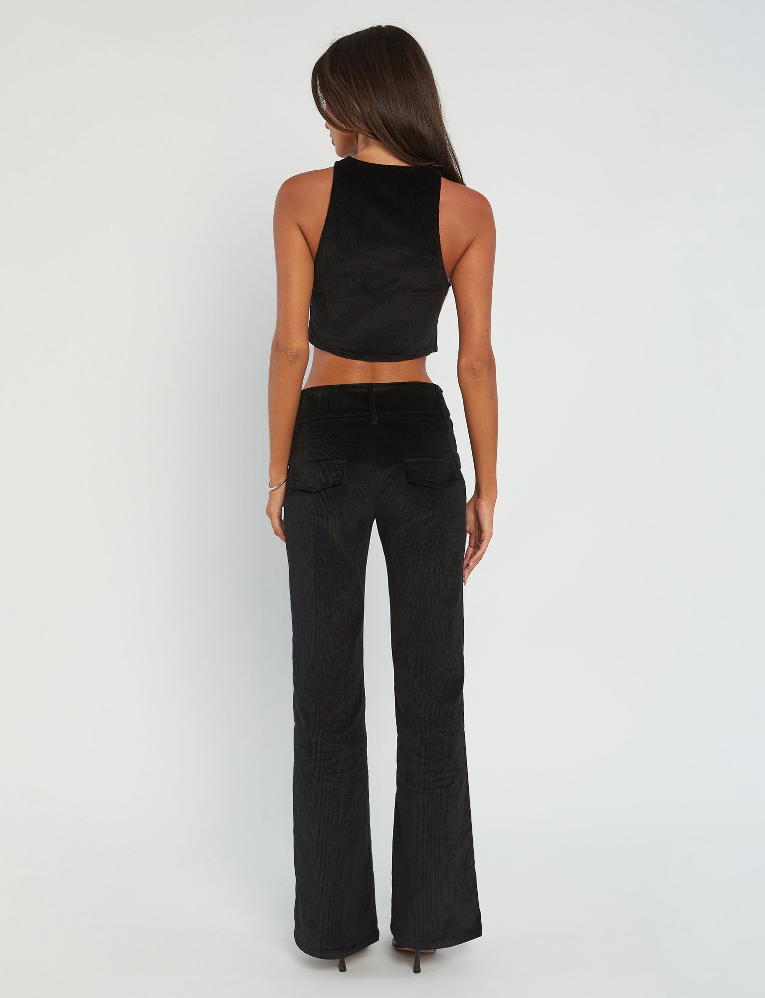 A woman with long dark hair wears a black sleeveless crop top and the ANEYA PANT - BLACK CORD, which features a low-rise waist and large square buckles. The flared silhouette accentuates her figure as she stands confidently in black sunglasses and open-toe heels against a plain white background.