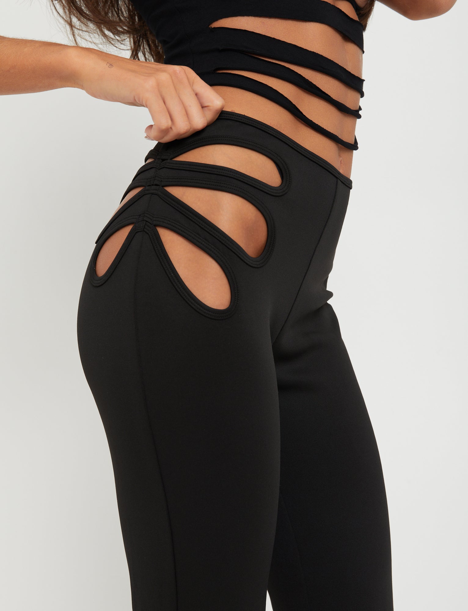 A person is wearing the KATO PANT - BLACK from TIGERMIST paired with a matching black top featuring multiple cut-out designs on the sides, reflecting Tiger Mist's edgy aesthetic. The image focuses on the waist and hip area, highlighting the unique style and cut-out details of the outfit. The background is plain white.