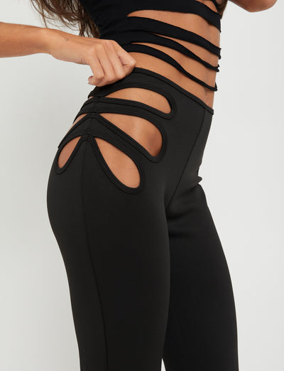 A person is wearing the KATO PANT - BLACK from TIGERMIST paired with a matching black top featuring multiple cut-out designs on the sides, reflecting Tiger Mist's edgy aesthetic. The image focuses on the waist and hip area, highlighting the unique style and cut-out details of the outfit. The background is plain white.