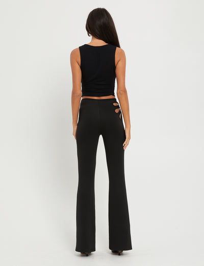 A woman with long dark hair stands with her back to the camera, wearing a sleeveless black crop top and TIGERMIST KATO PANT in black. The pants feature stylish cut-out details on one side near the waist. The background is plain white.