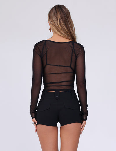 A woman with long blonde hair is seen from the back, wearing a black sheer long-sleeve top that reveals a black bra underneath and high-waisted KITTIE SHORT - BLACK from TIGERMIST. The shorts feature flap pockets with buttons. She is standing against a plain, light-colored background, sporting the best-selling KITTIE SHORT by TIGERMIST.