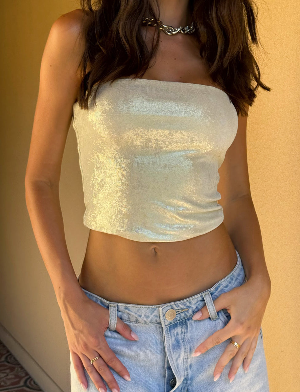 A person with long dark hair is wearing the COSIMA TOP - GOLD by TIGERMIST, paired with light blue high-waisted jeans. They have rings on their fingers and are posing with hands on their hips against a beige wall.