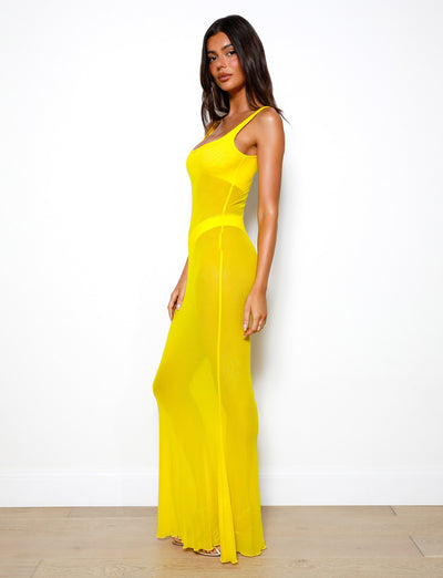 A woman stands against a plain white background, wearing the SAMIR DRESS - YELLOW from TIGERMIST. The sleeveless, long, sheer dress is a striking bright yellow. She has long, dark hair and gazes towards the camera with a neutral expression. The floor is wooden.