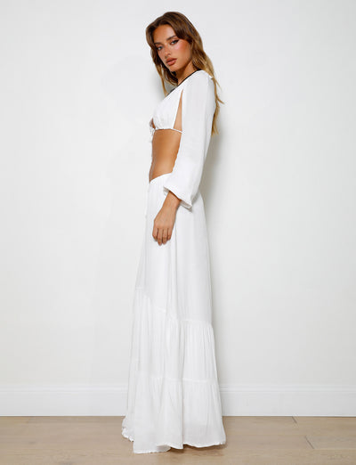 A woman stands against a plain white background, wearing the PHILLIPA SKIRT - WHITE by TIGERMIST with long sleeves and side cutouts at the waist. She is looking directly at the camera with a neutral expression, her blonde hair cascading over her shoulders.