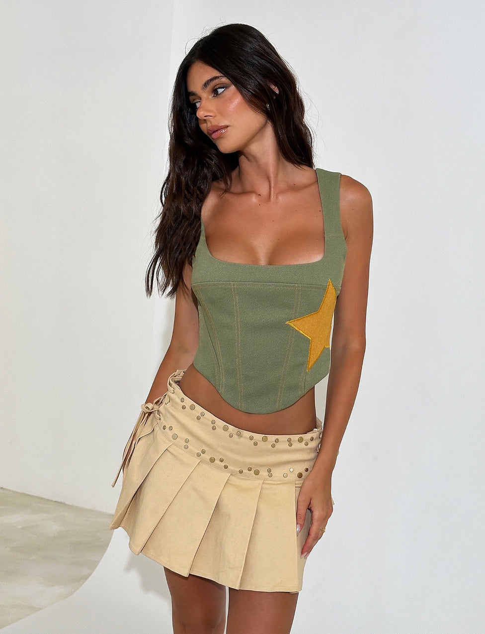 A woman with long, dark hair wears the SULLIVAN TOP - GREEN : KHAKI GREEN from TIGERMIST, featuring a large yellow star. She pairs it with a beige pleated mini skirt adorned with decorative studs from the same brand, standing against a plain white background and gazing off to the side.