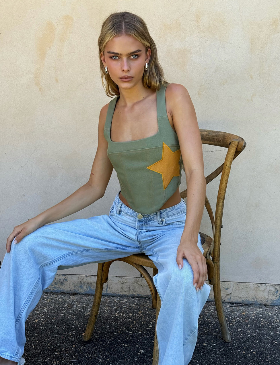 A person with medium-length, blonde hair is sitting on a wooden chair against a plain, light-colored wall. Clad in a stylish TIGERMIST SULLIVAN TOP in khaki green with a large yellow star on the side and loose, light blue jeans, they gaze directly at the camera with a neutral expression.