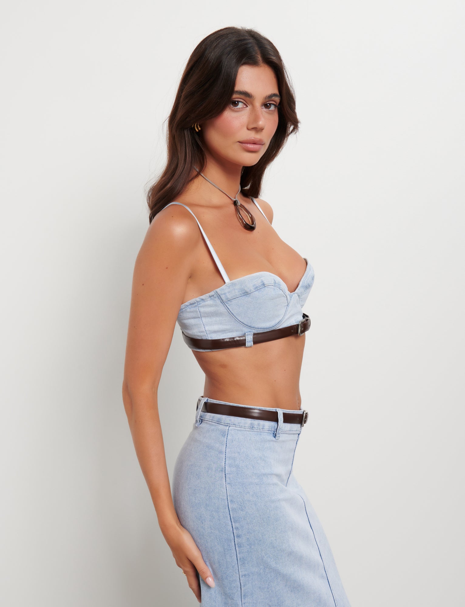 A woman with long dark hair is posing against a plain white background. She is wearing the light blue FLETCHER TOP - BLUE: DENIM BLUE by TIGERMIST with thin straps and a matching high-waisted denim skirt. She accessorizes with a brown belt and a matching brown pendant necklace.