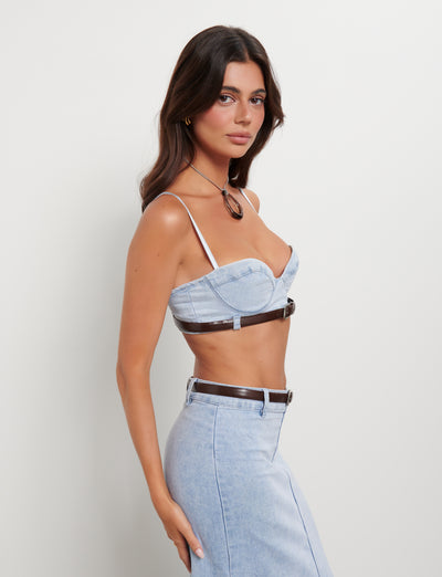 A woman with long dark hair is posing against a plain white background. She is wearing the light blue FLETCHER TOP - BLUE: DENIM BLUE by TIGERMIST with thin straps and a matching high-waisted denim skirt. She accessorizes with a brown belt and a matching brown pendant necklace.