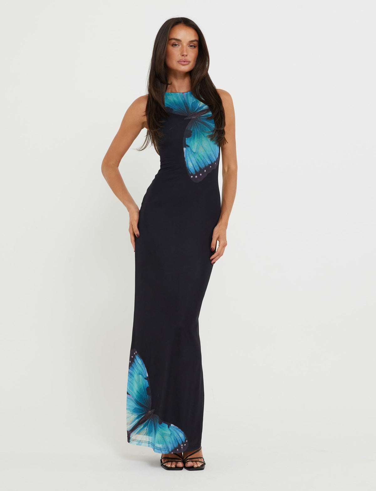 A woman with long dark hair stands against a plain white background, posing in the stylish ZAHRAA DRESS - BLACK : BUTTERFLY by TIGERMIST. The sleeveless, floor-length black dress features an eye-catching blue butterfly print on the chest and lower section. One hand is placed on her hip while the other rests by her side.