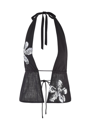 The MAISHA TOP from TIGERMIST is a black halter top with a deep V-neckline, featuring tie straps at the neck and back. It is adorned with sequined floral appliques on both sides, adding a touch of sparkle and elegance to the simple design.
