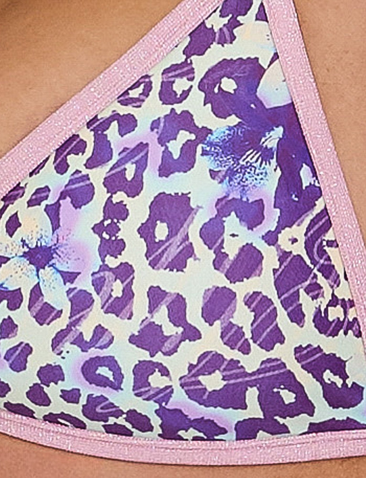 Close-up image of the TIGERMIST DIANA BIKINI TOP - BLUE : BY POPPY LEOPARD fabric featuring a purple and white leopard print pattern with pink trim. The design showcases an array of leopard spots in various shades of purple, complemented by white highlights and subtle light blue and white patches, creating an abstract visual effect.