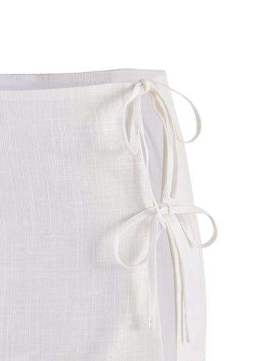 A close-up of the TIGERMIST NICOLA SKIRT - WHITE showcases two tied bows on the right side. The skirt's fabric features a textured pattern with subtle vertical and horizontal lines, creating a checkered appearance. The edges are neatly hemmed.