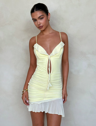 A woman wearing the CARLY DRESS - YELLOW : LEMON by TIGERMIST, a light yellow ruched mini dress with shoulder straps and a front tie, stands against a simple beige background. Her hair is pulled back, and she is accessorized with gold earrings and bracelets. She has a neutral expression with her hands by her sides.