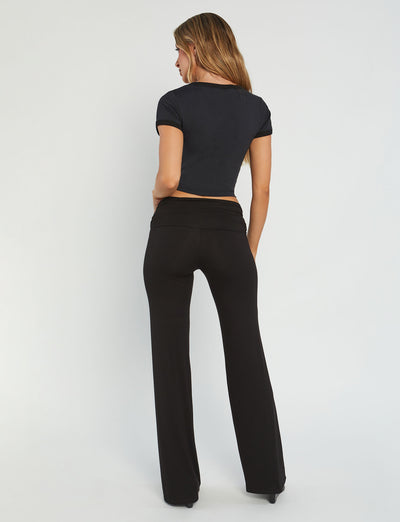 A woman stands with her back to the camera, dressed in the TIGERMIST BILLIE TEE - BLACK and matching black flared pants. Her long, wavy hair cascades down her back as she poses against a plain white backdrop.