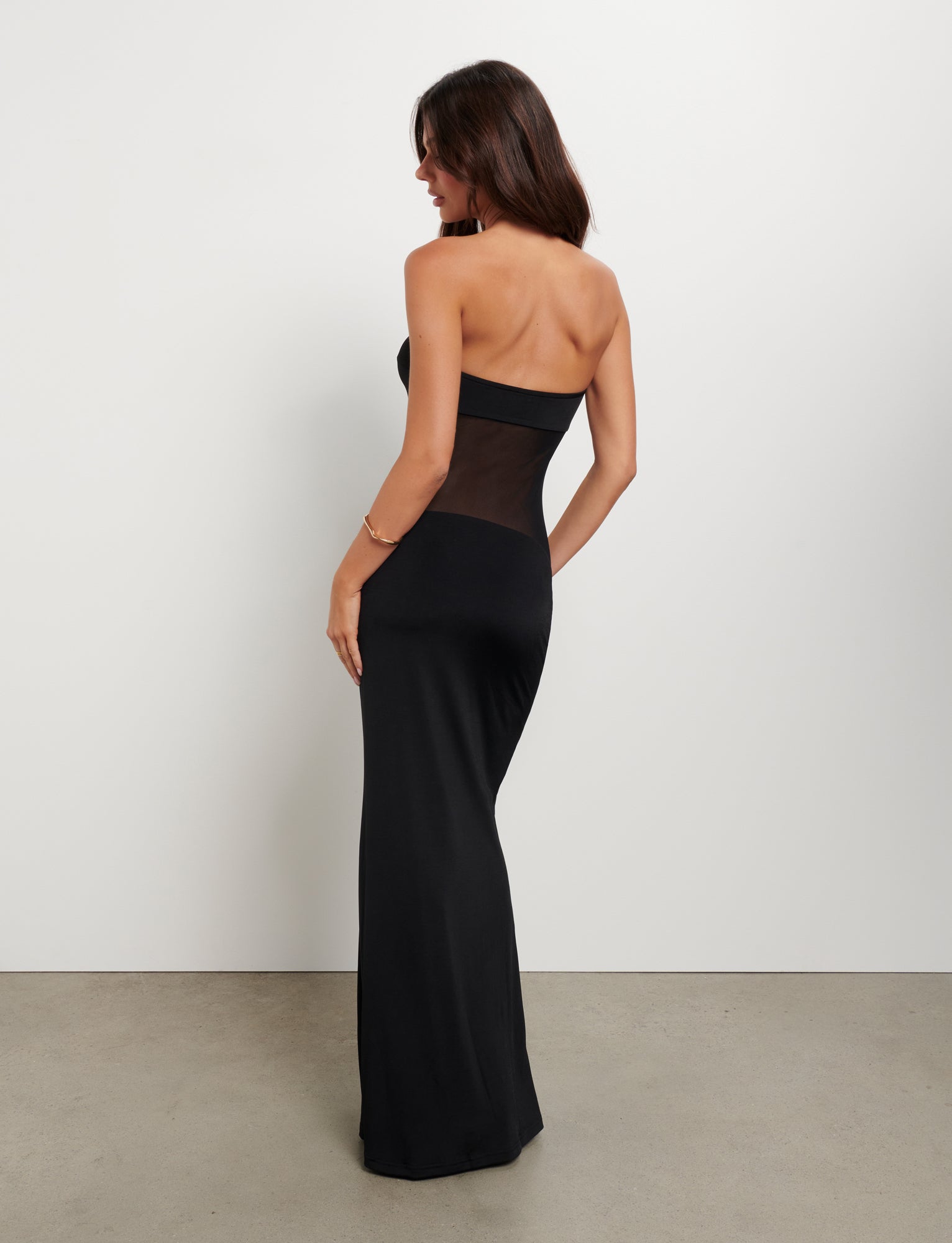 A woman with long dark hair stands facing away, highlighting the exquisite Tigermist Morgan Dress in black that beautifully accentuates her back. The strapless gown showcases a sheer panel along the back and cascades down to the floor. She accessorizes with a gold bracelet on one wrist against a plain white background.