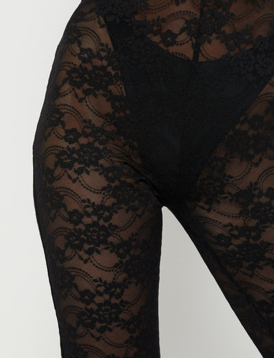 The image showcases a close-up of a person wearing the DEJA JUMPSUIT in black lace from TIGERMIST. The view is focused on the pant area, emphasizing the intricate floral lace design.