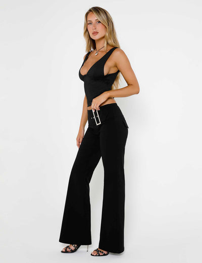 A woman with long blonde hair poses against a white background, confidently wearing the LUNARIA TOP in black and matching black flared pants from TIGERMIST. She holds a small black clutch and wears black high-heeled sandals, gazing assuredly towards the camera.