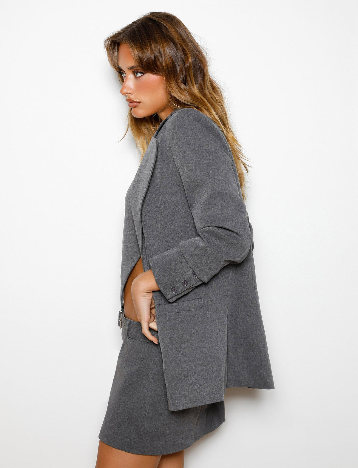 A person with long, wavy hair is standing against a plain white background, wearing the TIGERMIST NADINE JACKET - GREY : SLATE paired with matching shorts. They are facing sideways with a serious expression, accentuating the profile of their face and the relaxed fit of this Tiger Mist ensemble.