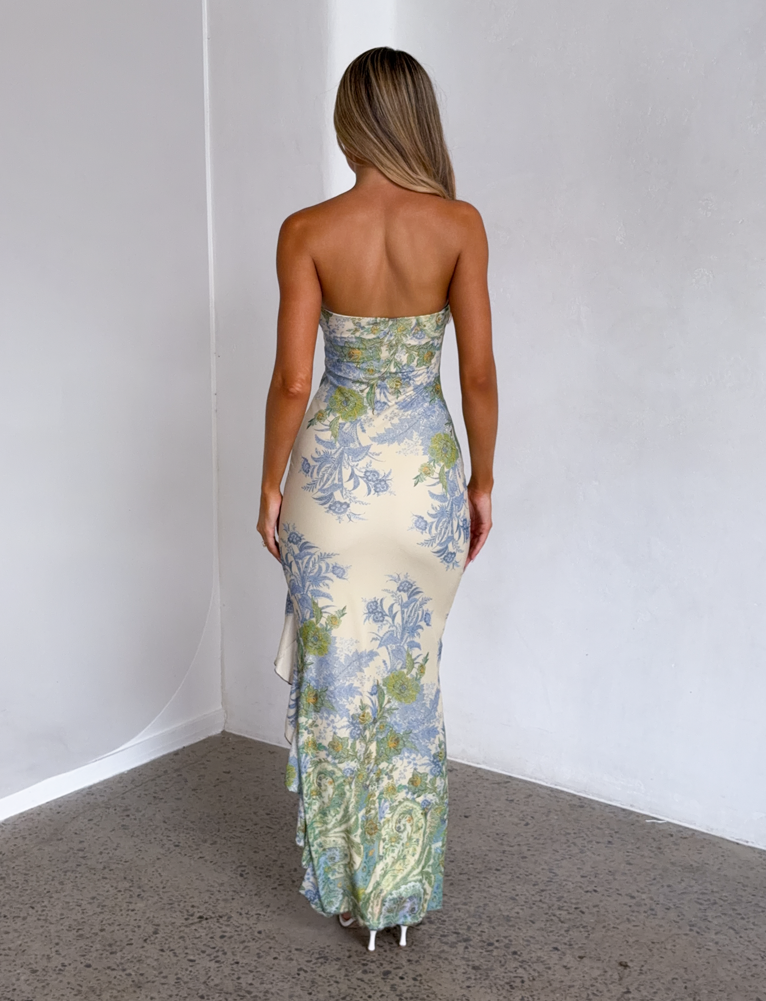 A woman with long, straight hair faces away from the camera, wearing the TIGERMIST THEO MAXI DRESS in multi paisley tapestry, featuring a blue and green floral pattern, in a minimalist white-walled room with a grey floor.
