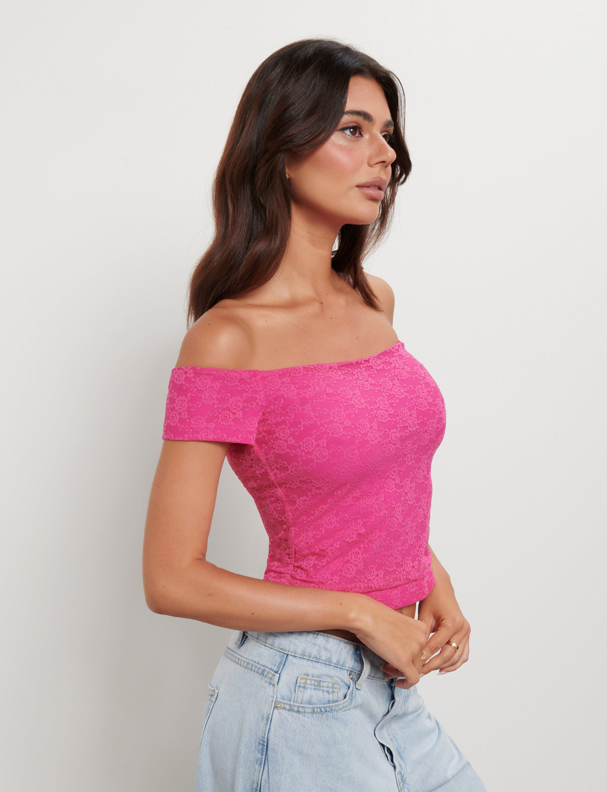 A young woman with long, dark hair is wearing the Caiden Top in hot pink lace from TIGERMIST paired with high-waisted light blue jeans. She stands against a plain white background, looking to the side with her hands loosely clasped in front of her.