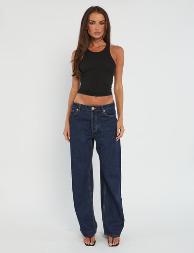 A woman stands against a plain white background, wearing the Tiger Mist SOMMER TANK in black and loose-fitting dark blue jeans. She has long, straight hair and is wearing black sandals. Her arms are relaxed by her sides.