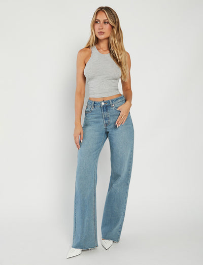 A woman with long, wavy blonde hair stands against a plain white background. Dressed in the SOMMER TANK - GREY and high-waisted blue jeans from TIGERMIST, she completes her look with white pointed-toe shoes. She has a relaxed expression, her left arm resting by her side while her right hand touches her hip.