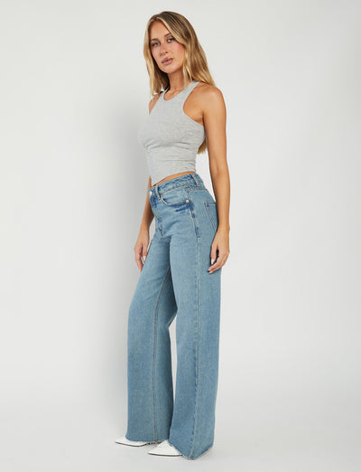 A woman with long hair wearing the SOMMER TANK - GREY sleeveless crop top and high-waisted, wide-leg blue jeans from TIGERMIST stands against a plain white background. She looks at the camera with a neutral expression, her right hand resting on her hip.