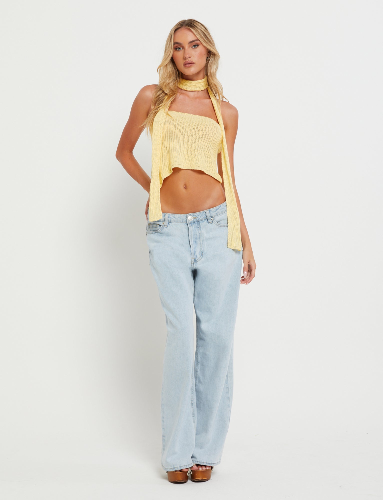 A woman stands against a plain white background wearing light blue high-waisted jeans from Tiger Mist, paired with the Lemon MORAI TOP by TIGERMIST, and a long, matching yellow scarf. She has long blonde hair and assumes a relaxed pose, with her hands by her sides and one foot slightly forward.