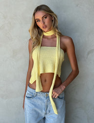 A person with long blonde hair stands in front of a gray textured wall. They are wearing the MORAI TOP - YELLOW : LEMON from TIGERMIST, paired with a matching long scarf and light blue jeans. With a serious expression and hands relaxed by their sides, they exude a cool confidence.
