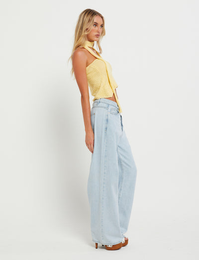 A woman with long blonde hair is posing against a plain white background. Clad in the sleeveless MORAI TOP - YELLOW : LEMON from TIGERMIST, paired with light blue wide-leg jeans, she stands in profile with her hands by her sides, looking off into the distance.