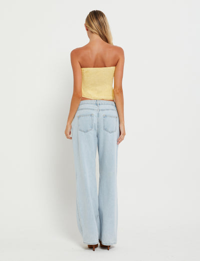 A woman with long blonde hair stands with her back to the camera, wearing a TIGERMIST MORAI TOP in Lemon and light blue high-waisted jeans against a plain white background.
